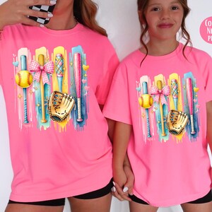 May include: Two pink t-shirts with a colorful graphic of baseball bats, a ball, a glove, and a bow. The text "Color: Neon Pink" is in the top right corner of the image.