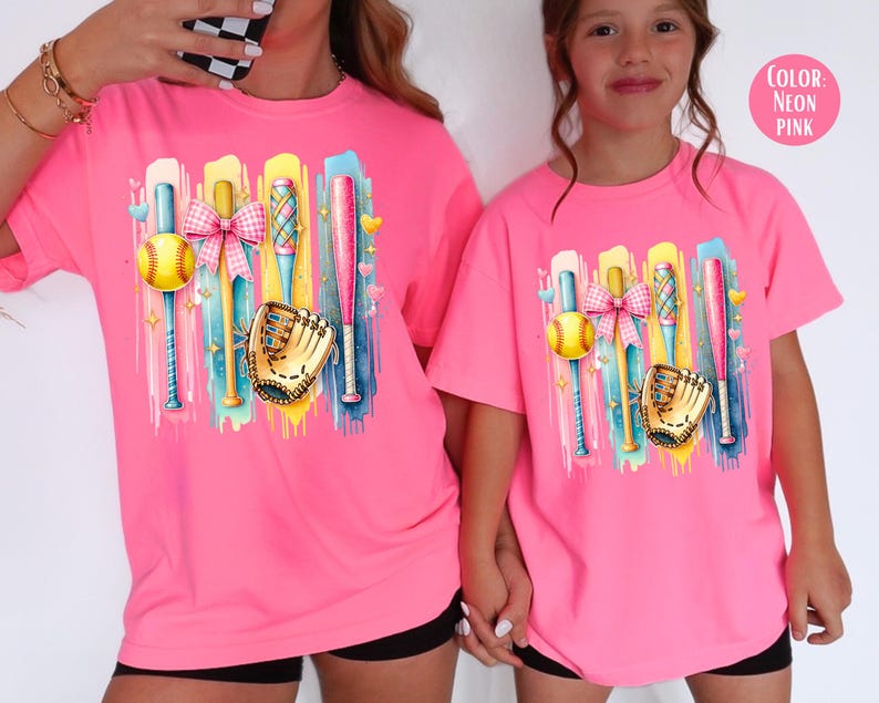 May include: Two pink t-shirts with a colorful graphic of baseball bats, a ball, a glove, and a bow. The text "Color: Neon Pink" is in the top right corner of the image.
