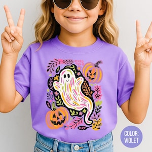May include: A violet t-shirt with a colourful ghost design, pumpkins, and floral accents. The ghost is white with colourful stripes. The pumpkins are orange with carved faces. The shirt has short sleeves.
