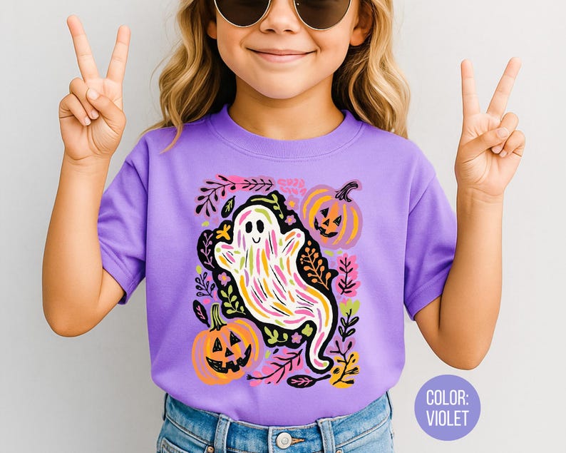 May include: A violet t-shirt with a colourful ghost design, pumpkins, and floral accents. The ghost is white with colourful stripes. The pumpkins are orange with carved faces. The shirt has short sleeves.