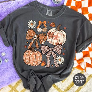 Peut inclure: T-shirt gris fonc&eacute; avec un motif sur le th&egrave;me de l'automne. Le design comprend des citrouilles, des n&oelig;uds et des &eacute;l&eacute;ments floraux dans des tons orange, marron et cr&egrave;me. La couleur du t-shirt est poivre.
