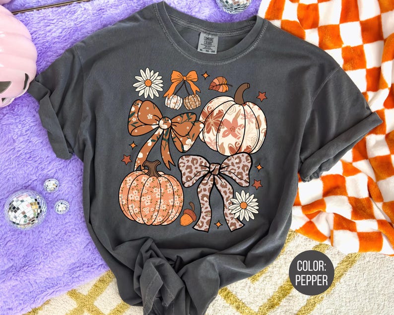 Peut inclure: T-shirt gris fonc&eacute; avec un motif sur le th&egrave;me de l'automne. Le design comprend des citrouilles, des n&oelig;uds et des &eacute;l&eacute;ments floraux dans des tons orange, marron et cr&egrave;me. La couleur du t-shirt est poivre.