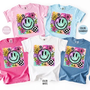 May include: A collection of short-sleeved t-shirts in various colors, including pink, blue, and white. Each shirt features a large, colorful graphic of a smiling face surrounded by flowers and a checkered pattern. The text "COLOR" and color names are visible.