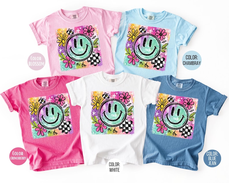 May include: A collection of short-sleeved t-shirts in various colors, including pink, blue, and white. Each shirt features a large, colorful graphic of a smiling face surrounded by flowers and a checkered pattern. The text "COLOR" and color names are visible.