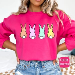 May include: A pink sweatshirt with a graphic of four pastel colored bunnies with their backs turned to the viewer. The bunnies are in a row and have white tails. The text "COLOR: HELICONIA" is in the bottom right corner of the image.