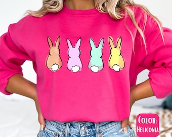 Könnte beinhalten: Ein rosa Sweatshirt mit einem Aufdruck von vier pastellfarbenen Hasen, die dem Betrachter den Rücken zukehren. Die Hasen stehen in einer Reihe und haben weiße Schwänze. Der Text "COLOR: HELICONIA" befindet sich in der rechten unteren Ecke des Bildes.