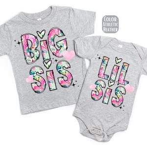 May include: Two gray toddler shirts, one a t-shirt and the other a onesie, with the words "Big Sis" and "Lil Sis" in a floral design. The text is outlined in black with pink, blue, and green floral patterns.