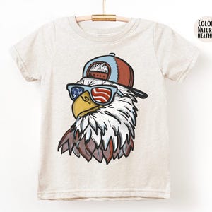 May include: A light beige t-shirt featuring a cartoon eagle wearing sunglasses with an American flag design and a red and blue baseball cap. The eagle has white and brown feathers. The shirt is hanging on a wooden hanger.