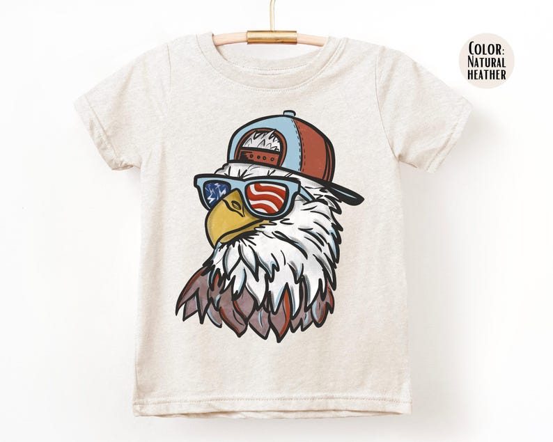 May include: A light beige t-shirt featuring a cartoon eagle wearing sunglasses with an American flag design and a red and blue baseball cap. The eagle has white and brown feathers. The shirt is hanging on a wooden hanger.