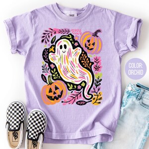 May include: Lilac t-shirt with a ghost and pumpkin design, surrounded by floral elements. The shirt is styled with checkered slip-on shoes and denim jeans. The text "COLOR: ORCHID" is displayed on the image.
