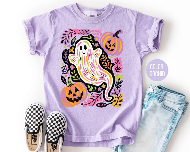 May include: Lilac t-shirt with a ghost and pumpkin design, surrounded by floral elements. The shirt is styled with checkered slip-on shoes and denim jeans. The text "COLOR: ORCHID" is displayed on the image.