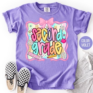 May include: A violet t-shirt with the words "Second Grade" in colorful bubble letters. The graphic includes a pink bow, flowers, stars, and a pencil. A pair of black and white checkered slip-on shoes and gray sweatpants are next to the shirt.