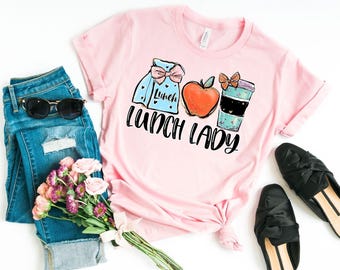 May include: Pink t-shirt with a graphic of a lunch bag, apple, and coffee cup. The text "Lunch Lady" is printed below the graphic.