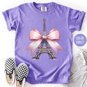 May include: Violet t-shirt featuring a graphic of the Eiffel Tower with a pink bow. The shirt has rolled sleeves and is paired with gray sweatpants and black and white checkered slip-on shoes. The text "COLOR: VIOLET" is in a circle.