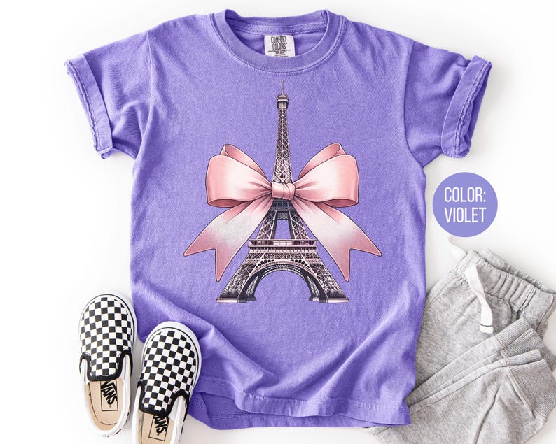 May include: Violet t-shirt featuring a graphic of the Eiffel Tower with a pink bow. The shirt has rolled sleeves and is paired with gray sweatpants and black and white checkered slip-on shoes. The text "COLOR: VIOLET" is in a circle.