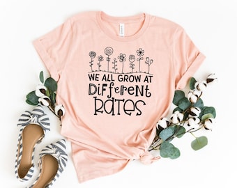 May include: A pink t-shirt with a black graphic that says "We all grow at different rates". The graphic also includes a row of stylized flowers.