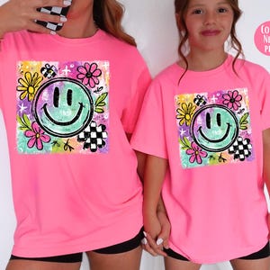 May include: Neon pink t-shirts featuring a colorful smiley face design. The graphic includes a green smiley face, flowers, and checkered elements. The shirts are worn by two people.