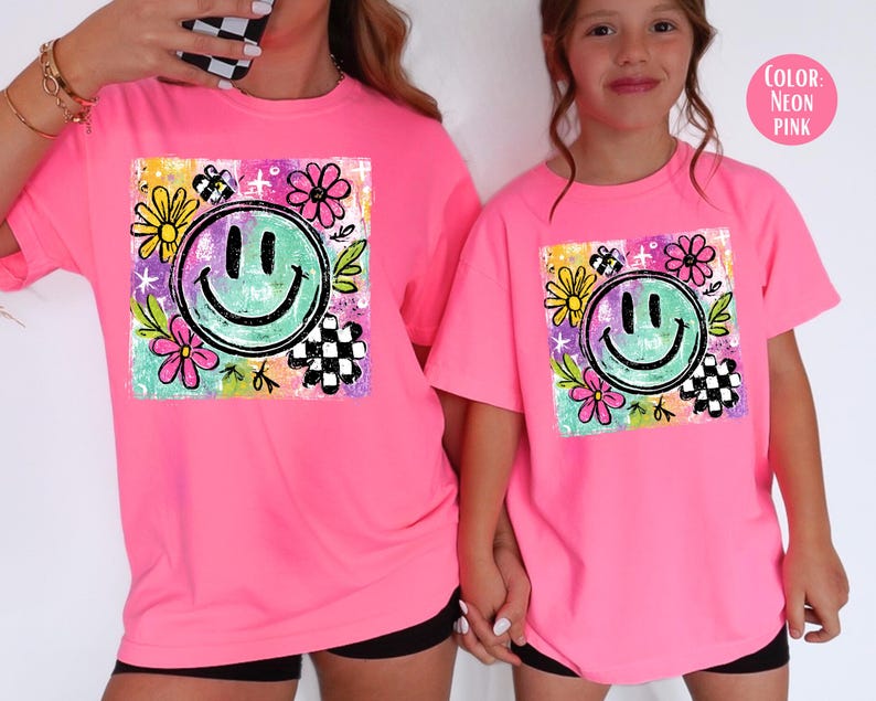 May include: Neon pink t-shirts featuring a colorful smiley face design. The graphic includes a green smiley face, flowers, and checkered elements. The shirts are worn by two people.