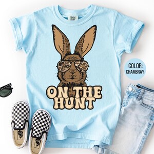 May include: Pale blue t-shirt with a brown rabbit wearing camouflage sunglasses. The words "ON THE HUNT" are printed below the rabbit. The image includes blue jeans, black sunglasses, and black and white checkered trainers.