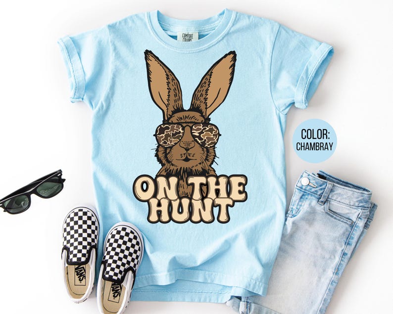 May include: Pale blue t-shirt with a brown rabbit wearing camouflage sunglasses. The words "ON THE HUNT" are printed below the rabbit. The image includes blue jeans, black sunglasses, and black and white checkered trainers.
