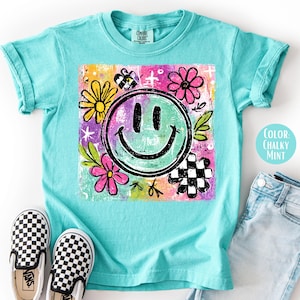 May include: A turquoise t-shirt with a smiley face graphic. The graphic includes a green smiley face, flowers, and a checkered pattern. The text "Color: Chalky Mint" is visible on the shirt. Black and white checkered slip-on shoes are next to the shirt.