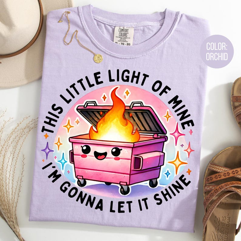 May include: A purple t-shirt featuring a cartoon image of a pink dumpster on fire with the text "This little light of mine I'm gonna let it shine".