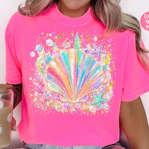 May include: Neon pink t-shirt featuring a colorful seashell design with starfish and aquatic elements. The text "COLOR: NEON PINK" is in a pink circle. The shirt is a vibrant, eye-catching piece.