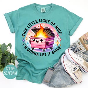 May include: A teal blue t-shirt featuring a cartoon image of a pink bin with a smiling face and flames coming out of it. The text "This little light of mine I'm gonna let it shine" is printed around the image.