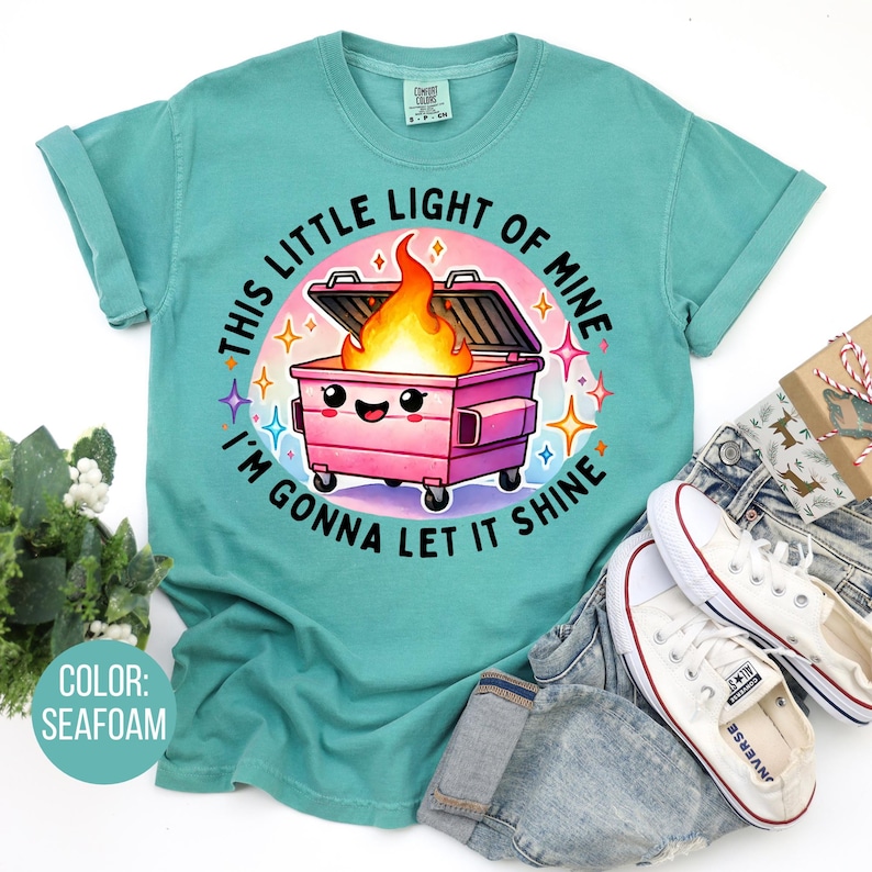 May include: A teal blue t-shirt featuring a cartoon image of a pink bin with a smiling face and flames coming out of it. The text "This little light of mine I'm gonna let it shine" is printed around the image.