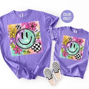 May include: Two violet t-shirts, one adult and one child size, featuring a colorful graphic. The graphic includes a smiling face, flowers, and a checkered pattern. A pair of black and white checkered slip-on shoes are next to the child's shirt.