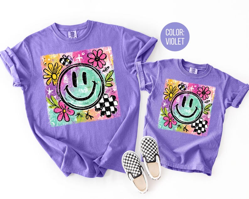 May include: Two violet t-shirts, one adult and one child size, featuring a colorful graphic. The graphic includes a smiling face, flowers, and a checkered pattern. A pair of black and white checkered slip-on shoes are next to the child's shirt.