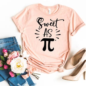 May include: A peach-colored t-shirt with the text "Sweet as Pi" in black lettering. The shirt is laid flat on a white surface with a pair of light brown high heels and a pair of blue jeans with pink flowers.