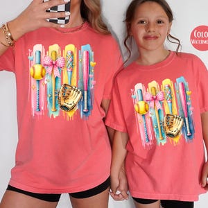 May include: Two t-shirts with a colorful graphic of baseball bats, a softball, a glove, and a pink bow. The t-shirts are a watermelon color.