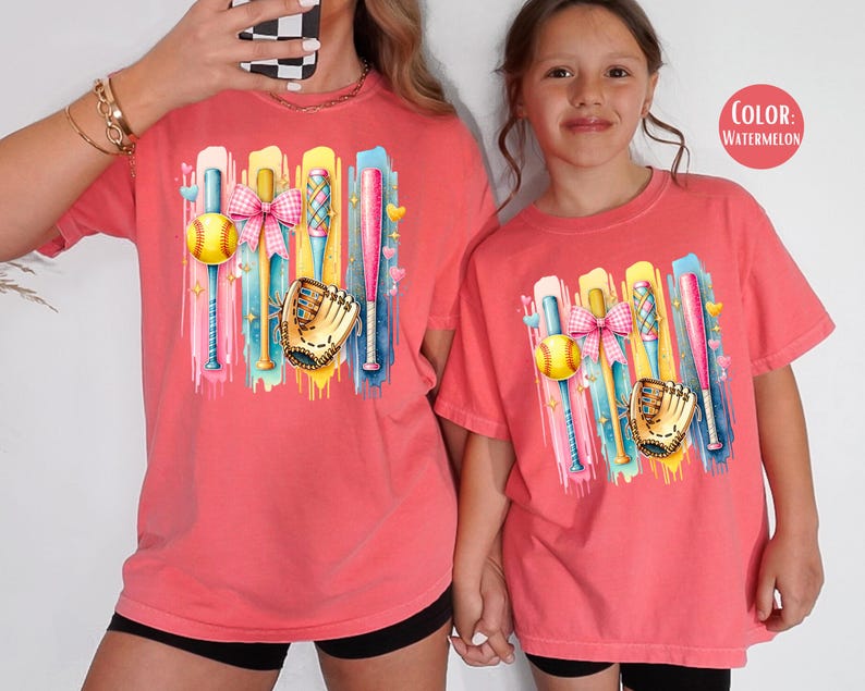 May include: Two t-shirts with a colorful graphic of baseball bats, a softball, a glove, and a pink bow. The t-shirts are a watermelon color.