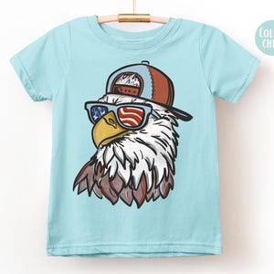 May include: A light blue t-shirt featuring a cartoon eagle wearing sunglasses with an American flag design and a brown and blue baseball cap. The eagle has white and brown feathers and a yellow beak. The shirt is hanging on a wooden hanger.