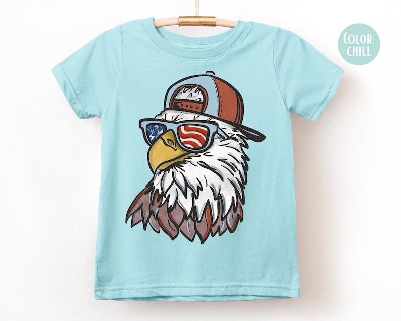 May include: A light blue t-shirt featuring a cartoon eagle wearing sunglasses with an American flag design and a brown and blue baseball cap. The eagle has white and brown feathers and a yellow beak. The shirt is hanging on a wooden hanger.