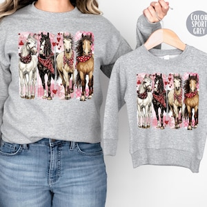May include: Sport grey sweatshirt featuring a graphic of four horses with red bandanas, set against a pink and white background with hearts. The sweatshirt is shown in adult and child sizes.