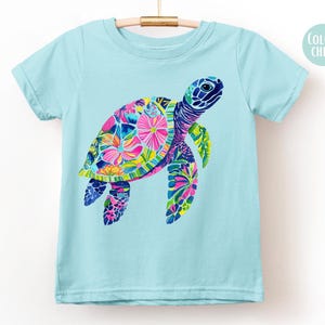 May include: A light blue t-shirt featuring a colorful sea turtle design. The turtle's shell is adorned with vibrant pink, yellow, and blue floral patterns. The shirt is hanging on a wooden hanger.