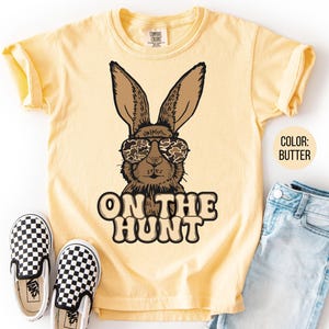 May include: A butter-yellow t-shirt with a brown rabbit wearing camouflage sunglasses. The text "ON THE HUNT" is printed below the rabbit. The shirt is styled with blue jeans and black and white checkered slip-on shoes.
