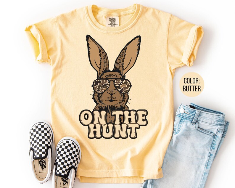 May include: A butter-yellow t-shirt with a brown rabbit wearing camouflage sunglasses. The text "ON THE HUNT" is printed below the rabbit. The shirt is styled with blue jeans and black and white checkered slip-on shoes.