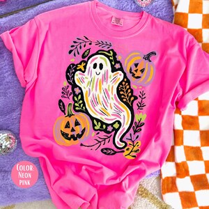 May include: Bright neon pink t-shirt with a Halloween-themed design. The graphic features a smiling ghost with colorful stripes, pumpkins, and foliage. The shirt's design includes two jack-o'-lanterns and black leaves. A vibrant and eye-catching garment.