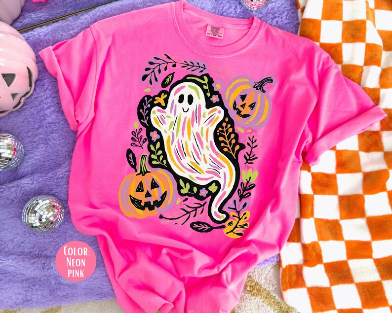 May include: Bright neon pink t-shirt with a Halloween-themed design. The graphic features a smiling ghost with colorful stripes, pumpkins, and foliage. The shirt's design includes two jack-o'-lanterns and black leaves. A vibrant and eye-catching garment.