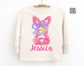 May include: A long-sleeved, natural heather-colored shirt featuring a colorful bunny design. The bunny is depicted in shades of pink, blue, and yellow, with a heart and bow. The name "Jessica" is written in pink script below the bunny.
