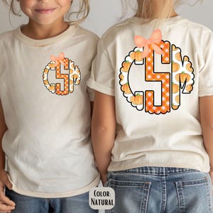 May include: Two children wearing natural-colored t-shirts with a pumpkin-themed monogram design. The monogram features the letter "S" in orange gingham, surrounded by pumpkins and a peach bow. The shirts are paired with blue jeans.