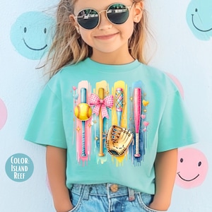 May include: A turquoise t-shirt with a colorful graphic of baseball bats, a softball, a glove, and a pink bow. The graphic is on a light blue background with white paint splatters.