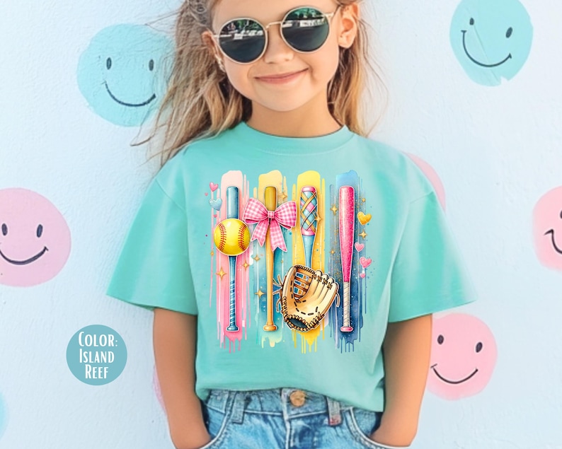 May include: A turquoise t-shirt with a colorful graphic of baseball bats, a softball, a glove, and a pink bow. The graphic is on a light blue background with white paint splatters.
