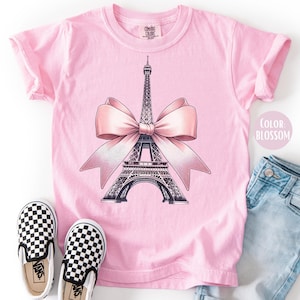 May include: Pink t-shirt featuring a graphic of the Eiffel Tower with a pink bow. The shirt is paired with checkered slip-on shoes and light blue denim jeans. The text "Color Blossom" is visible.