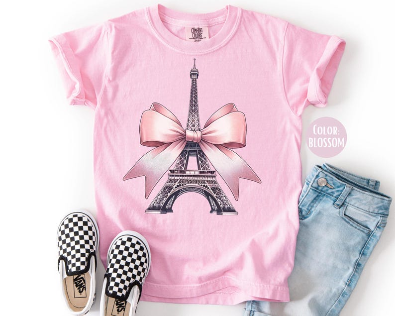 May include: Pink t-shirt featuring a graphic of the Eiffel Tower with a pink bow. The shirt is paired with checkered slip-on shoes and light blue denim jeans. The text "Color Blossom" is visible.