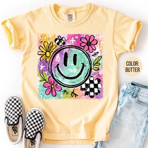 May include: Butter-colored t-shirt with a colorful graphic featuring a smiling face, flowers, and a checkered pattern. The shirt is paired with blue jeans and black and white checkered slip-on shoes.