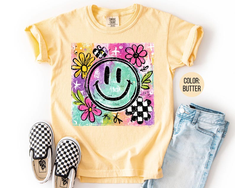 May include: Butter-colored t-shirt with a colorful graphic featuring a smiling face, flowers, and a checkered pattern. The shirt is paired with blue jeans and black and white checkered slip-on shoes.